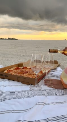 two glasses of wine are sitting on a towel near a pizza box and the ocean