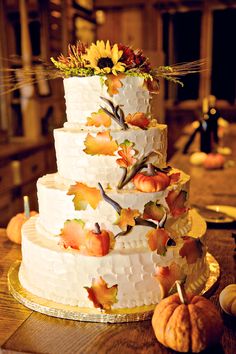 a three tiered white wedding cake decorated with fall leaves and sunflowers on a wooden table