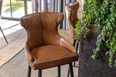 a brown leather bar stool next to a potted plant in front of a window