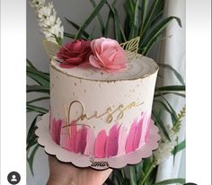 a hand holding a cake with pink frosting and flowers on top, in front of a plant