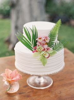 a white wedding cake with flowers and greenery on top, sitting on a wooden table