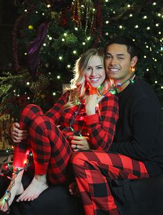 a man and woman dressed in plaid pajamas sitting next to a christmas tree with lights