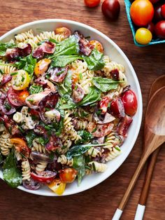 a white bowl filled with pasta salad next to wooden spoons and tomatoes on the table