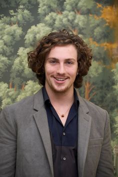 a man with curly hair wearing a gray jacket and black shirt smiling at the camera