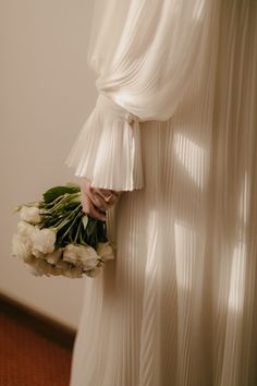 a woman in a white dress holding a bouquet of flowers behind her back with sheer curtains