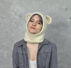 a woman wearing a knitted bear hat and scarf over her head, standing in front of a concrete wall