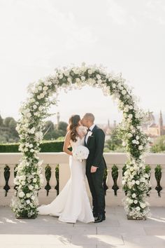 a bride and groom kissing in front of an arch decorated with white flowers at their wedding