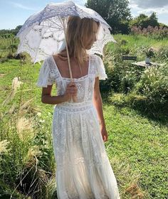 a woman in a white dress is holding an umbrella and standing on the grass with her back to the camera