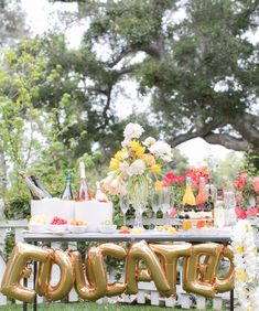 a table topped with gold balloons that spell out the word happy in front of flowers and greenery