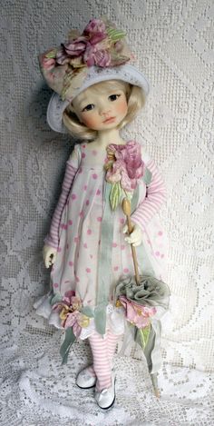 a doll with a hat and dress holding a flower in her hand on a white lace background