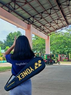 a woman in blue shirt carrying a black and yellow bag under a covered area with trees