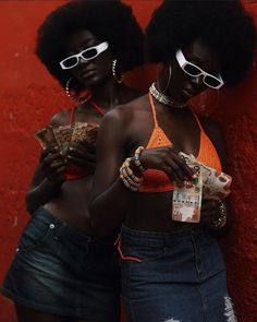 two black women standing next to each other in front of a red wall with sunglasses on