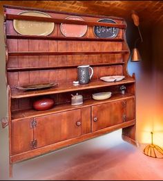 an old fashioned wooden dresser with plates and cups on it's shelf, next to a lamp