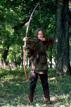 a woman dressed in medieval clothing is holding a bow and arrow while standing in the woods