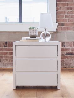 a white dresser with a lamp on top of it in front of a brick wall