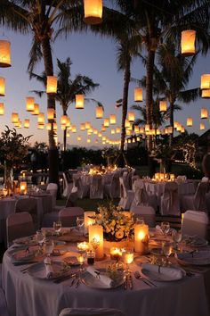 an outdoor dining area with lit candles and paper lanterns in the sky above it, surrounded by palm trees