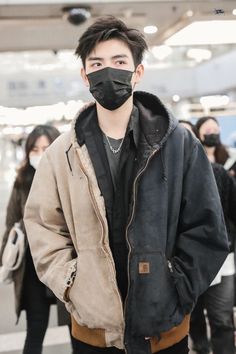 a man wearing a black face mask and jacket at an airport with people in the background