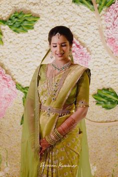 a woman in a green and gold sari with jewelry on her neck, standing next to a floral wall