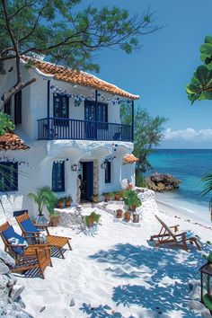 a white house with blue shutters on the beach next to trees and chairs in front of it