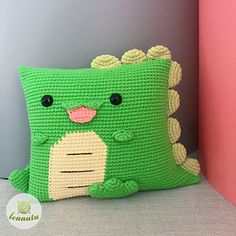 a green crocheted pillow with a smiling face on it's side, sitting next to a pink wall