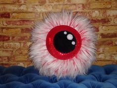 a red and white stuffed animal eyeball sitting on top of a blue cushion in front of a brick wall