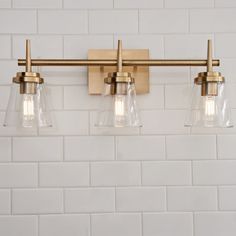 three light bathroom fixture with clear glass shades on white tile wall and gold metal fixtures