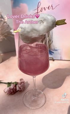 a pink drink in a wine glass with cotton floss on top and the words lover drinks the archer above it