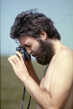 a shirtless man holding a camera up to his face while standing in the grass