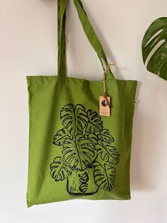 Eco-Friendly recycled plastic RPET Monstera Deliciosa tote bag. The tote bag is a lushes cardamon green colour and features a black linocut design of the popular house plant Monstera Deliciosa or Swiss cheese plant.  Made from recycled plastic bottles, this bag is a stylish and sustainable way to carry your belongings. This stylish lightweight RPET Luxury Shopper bag is made from recycled plastic which has been diverted from ending up in the ocean or landfill. A timeless classic integrated with ethical production. RPET Tote Bags are the promotion to a circular way. The bag is the perfect size for use as a luxury shopper or everyday tote, with ample space to carry all of your essentials, Whether you're heading to the beach, running errands, or simply out and about, the durable RPET material Organic Recyclable Tote Bag, Eco-friendly Recyclable Tote Shoulder Bag, Casual Green Reusable Bag, Eco-friendly Recyclable Shoulder Bag, Green Tote Gift Bag, Eco-friendly Green Recyclable Shoulder Bag, Casual Green Recyclable Bags, Green Recyclable Shoulder Bag For Everyday, Eco-friendly Green Bags For Gifts