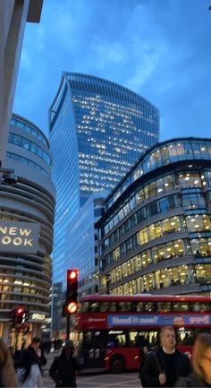 people are crossing the street in front of some tall buildings at dusk, with one red bus passing by