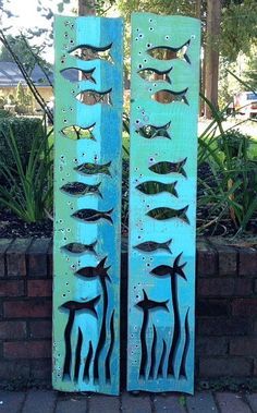 two metal fish sculptures sitting next to each other on a brick wall in front of a garden