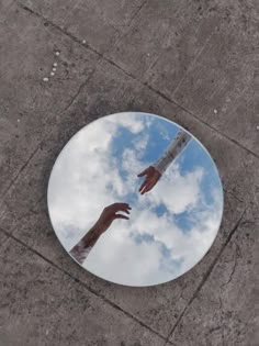 two hands reaching for each other in front of a round mirror on the ground with sky and clouds reflected in it