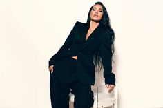 a woman in a black suit leaning against a white wall and posing for the camera