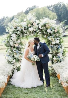 a bride and groom kissing in front of an arch with flowers on the grass at their wedding