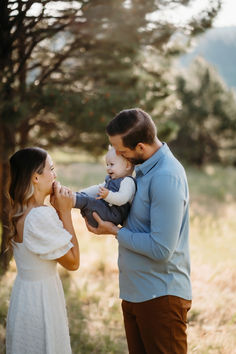 a man holding a baby while standing next to a woman in a field with trees