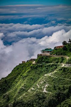 the top of a green mountain with clouds in the sky and buildings at the top