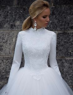 a woman wearing a white wedding dress with long sleeves and a high neckline is standing in front of a stone wall