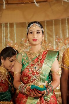 a woman in a red and green sari standing next to another woman wearing gold jewelry