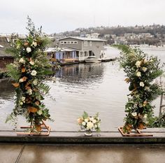 flowers and greenery are arranged on the edge of a dock with boats in the background