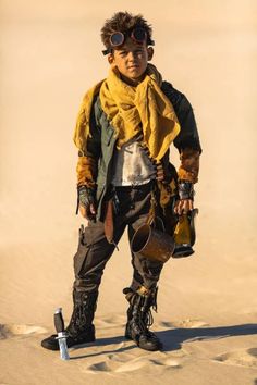 a young boy dressed in steampunk clothing and goggles, standing in the sand