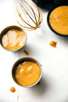 three metal cups filled with orange sauce on top of a white table next to other bowls