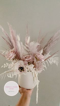 a hand holding a white vase with pink flowers and feathers on it's side