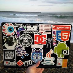 someone is holding up a laptop with stickers on it near the water and beach