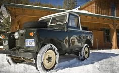 an old green truck parked in front of a log cabin with snow on the ground