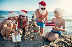 three kids and an adult in santa hats are on the beach near a christmas tree