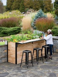 a woman standing at an outdoor bar with plants growing on it and stools next to it