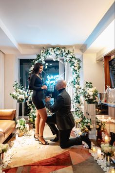 a man kneeling down next to a woman in front of a wedding arch with flowers on it