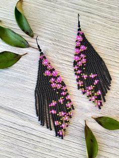 two black and pink beaded earrings sitting on top of a wooden table next to green leaves