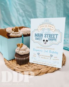 cupcakes are sitting on a wicker tray next to a sign that says join us for a main street sweet shoppe