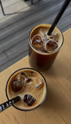 two glasses filled with liquid and ice on top of a wooden table next to each other